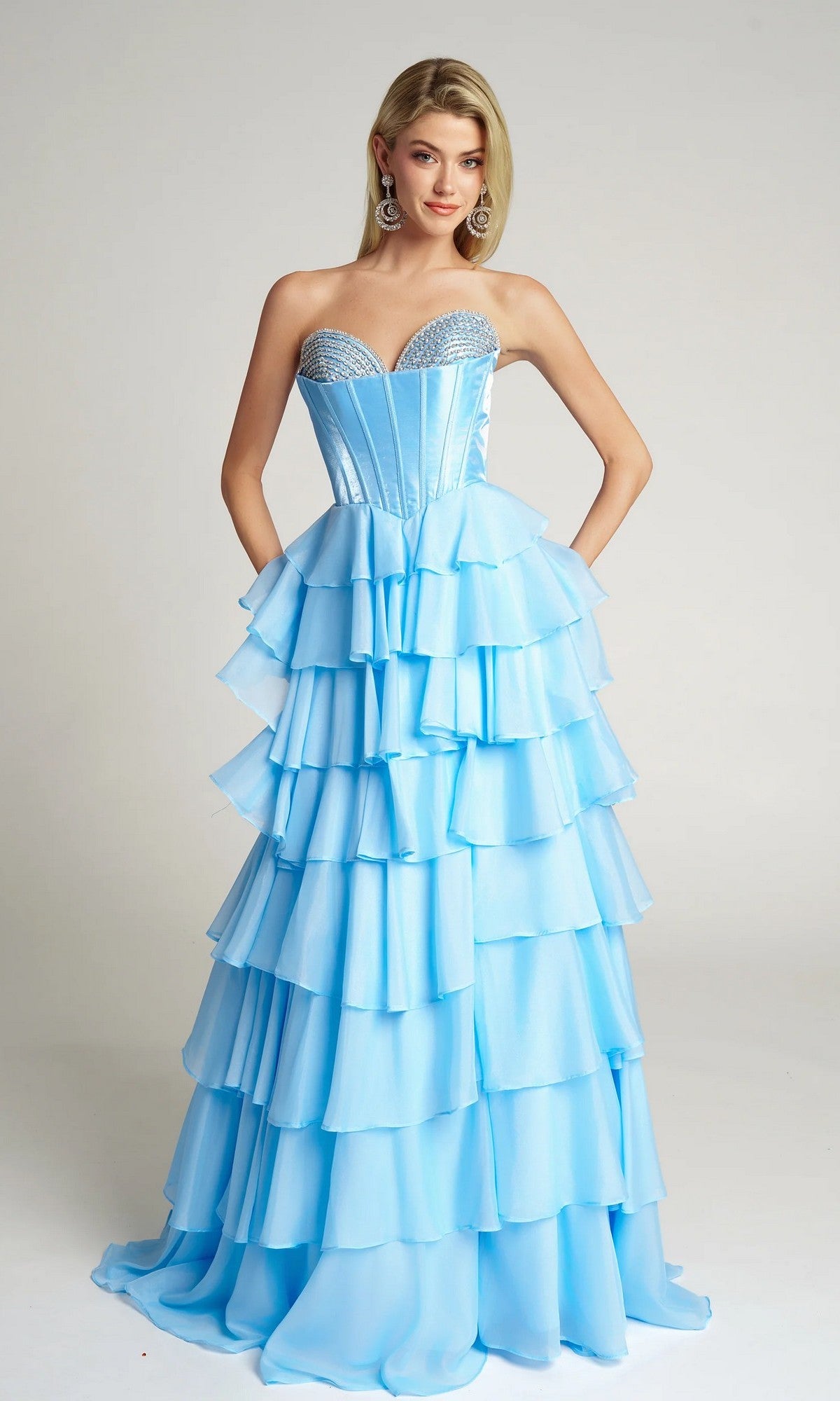 A woman models the blue Portia and Scarlett PS26024 long prom dress, featuring a strapless velvet corset bodice and tiered ruffle A-line skirt, standing against a plain background.